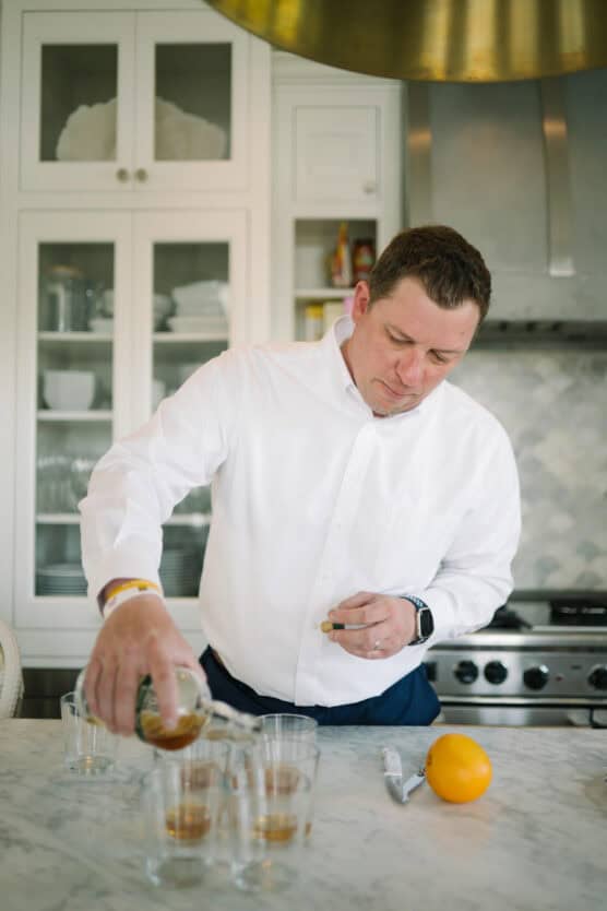 A man in a white shirt pours a drink into glasses on a marble kitchen counter. An orange and a peeler sit nearby. Cabinets with dishes and a stove are visible in the background. by Justin Salem Meyer