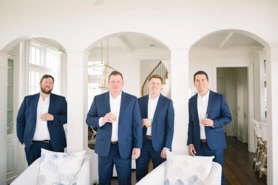 Four men wearing blue suits and white shirts stand indoors, smiling and holding drinks. They are positioned in front of white arches in a bright, elegant room with modern decor. by Justin Salem Meyer