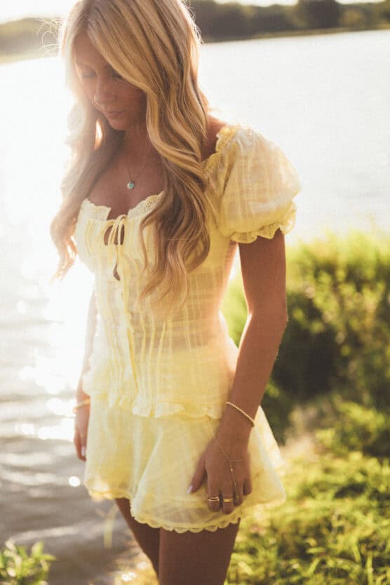 A woman with long blonde hair wearing a light yellow, short-sleeved dress stands near a sunlit body of water, with greenery in the background. She looks down, and the scene is softly lit. by Justin Salem Meyer