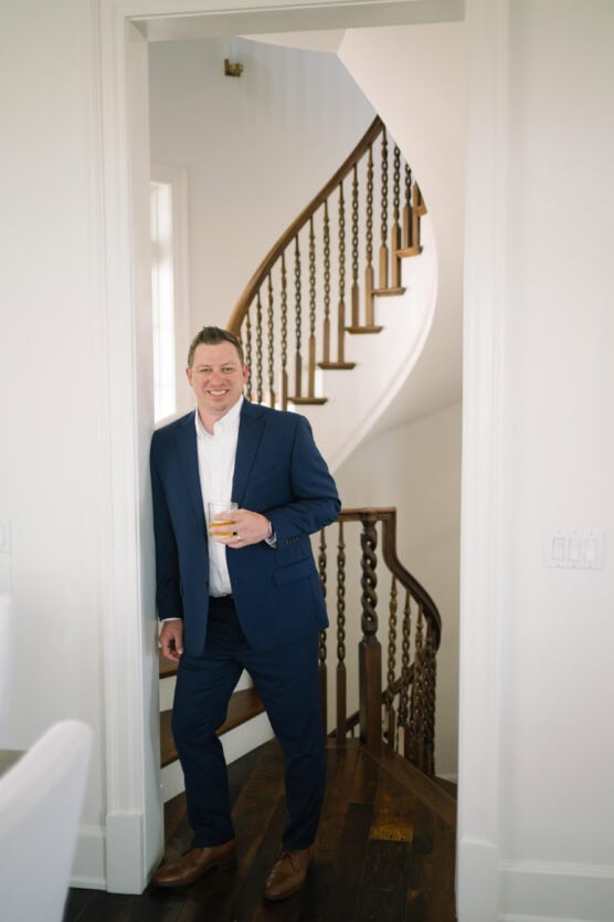 A man in a blue suit holding a drink stands smiling in a doorway with a spiral wooden staircase in the background. The setting appears to be indoors with bright, natural light. by Justin Salem Meyer