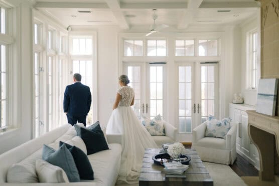 A bride in a white gown with lace details stands behind a man in a suit, both facing large, bright windows in a sunlit, elegant living room with white furniture and blue accents. by Justin Salem Meyer
