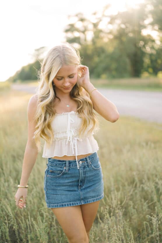 A young woman with long blonde hair walks through a field, wearing a white sleeveless top and a denim skirt, with sunlight softly illuminating the background. She looks down and gently touches her hair, appearing relaxed and content. by Justin Salem Meyer