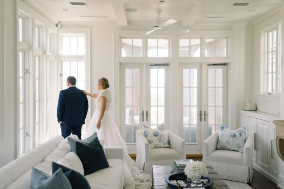 A bride in a white gown gently touches the shoulder of a man in a suit as they stand together by tall, sunlit windows in a bright, elegant living room with white furniture and blue accents. by Justin Salem Meyer