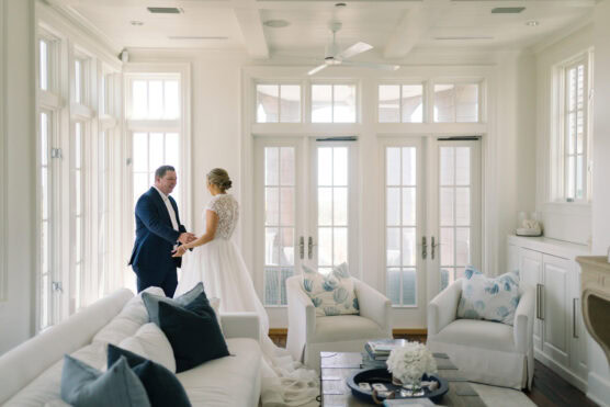 A bride and groom stand holding hands in a bright, elegant living room with large windows, white walls, and cozy white furniture accented with blue and gray pillows. by Justin Salem Meyer