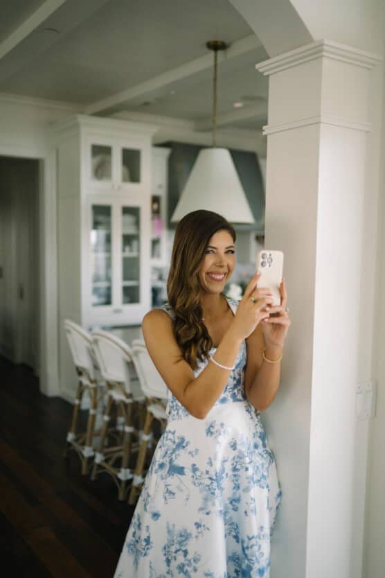A woman in a blue and white floral dress stands indoors, smiling and taking a selfie with her phone in a bright, modern kitchen. by Justin Salem Meyer