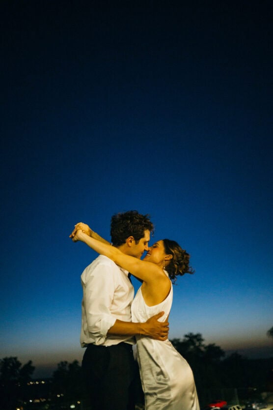 A couple embraces and smiles at each other under a deep blue twilight sky, with trees and lights faintly visible in the background. The woman wears a white dress and the man wears a white shirt and dark pants. by Justin Salem Meyer
