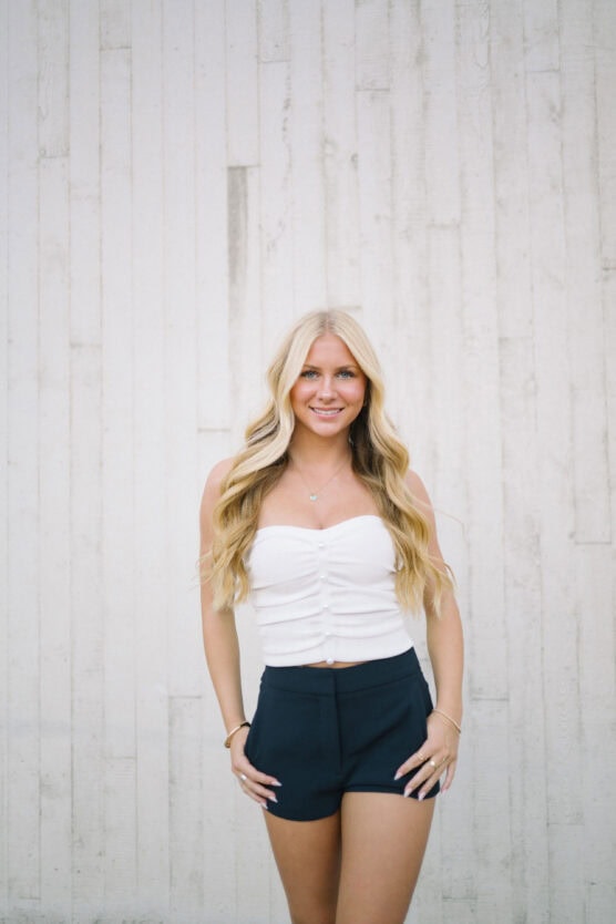 A young woman with long blonde hair, wearing a white strapless top and black shorts, stands smiling against a light-colored, vertically paneled wall. by Justin Salem Meyer