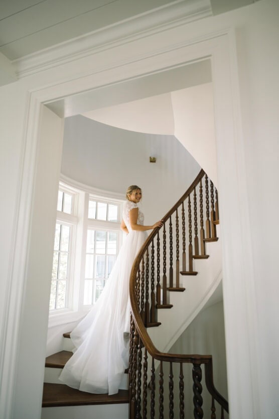 A bride in a white dress stands gracefully on a wooden spiral staircase, looking over her shoulder. Sunlight streams through large windows, illuminating her and the elegant interior. by Justin Salem Meyer