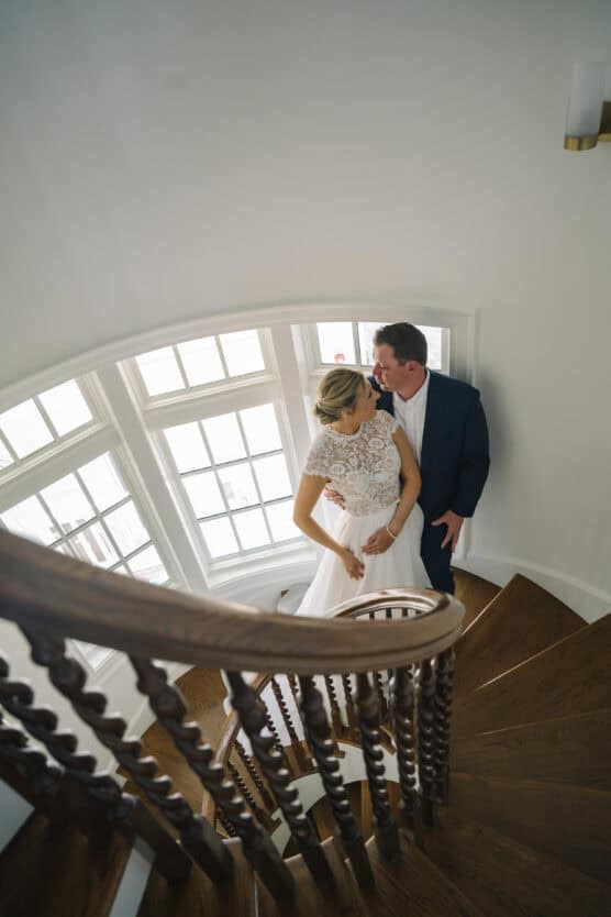 A bride and groom stand close together on a wooden spiral staircase, gazing at each other lovingly, with large bright windows behind them illuminating the scene. by Justin Salem Meyer