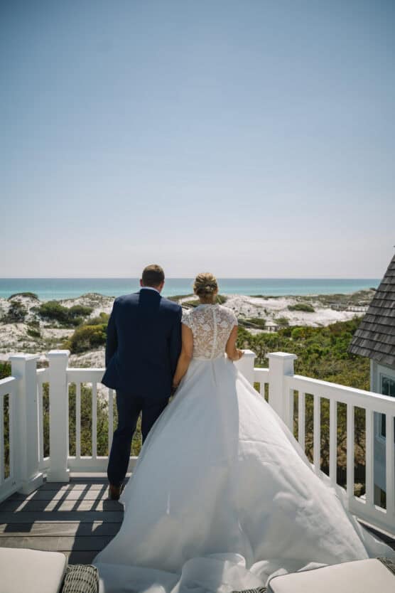 A bride and groom stand side by side on a white balcony, facing the ocean and sandy dunes under a clear blue sky, holding hands and wearing formal wedding attire. by Justin Salem Meyer