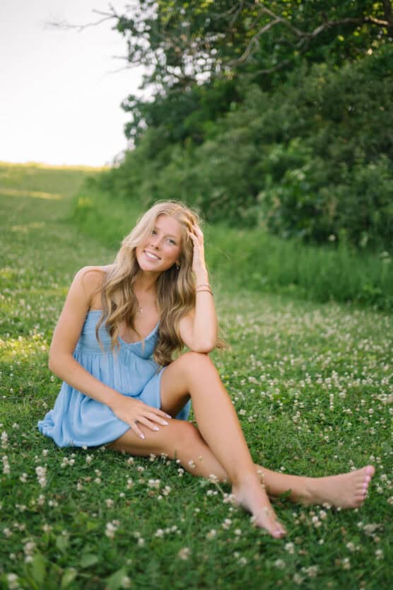 A young woman with long blonde hair sits barefoot on grass, wearing a light blue dress. She smiles at the camera, with one hand touching her hair. Green trees and clover flowers surround her on a sunny day. by Justin Salem Meyer