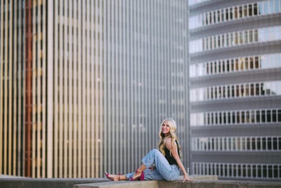 A woman with long blonde hair sits on a ledge, smiling, wearing a black top, light blue jeans, and pink shoes. Tall office buildings with many windows are in the background. by Justin Salem Meyer