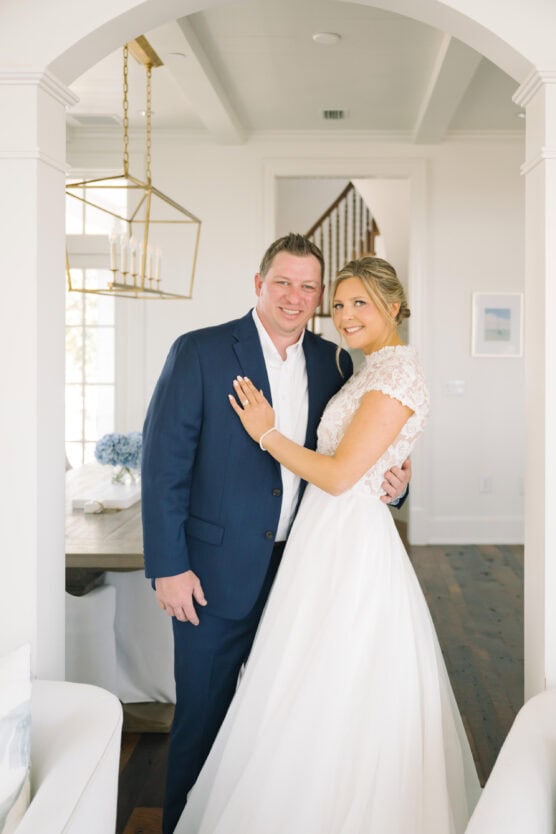 A smiling couple poses indoors; the man wears a navy suit and the woman wears a white lace wedding dress. They stand together in a bright, elegant room with white walls and wooden floors. by Justin Salem Meyer