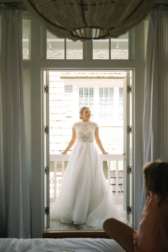 A bride in a white dress stands on a balcony framed by open doors, looking serene. A young girl sits on a bed in the foreground, gazing toward the bride. Soft light fills the airy room. by Justin Salem Meyer