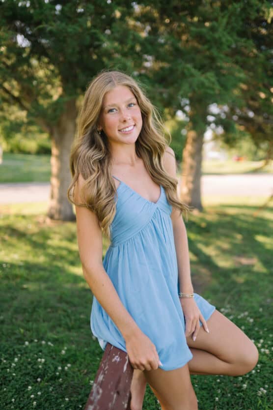 A young woman with long, wavy blonde hair wearing a light blue dress stands outdoors in a grassy area, leaning against a wooden bench with trees and sunlight in the background. by Justin Salem Meyer
