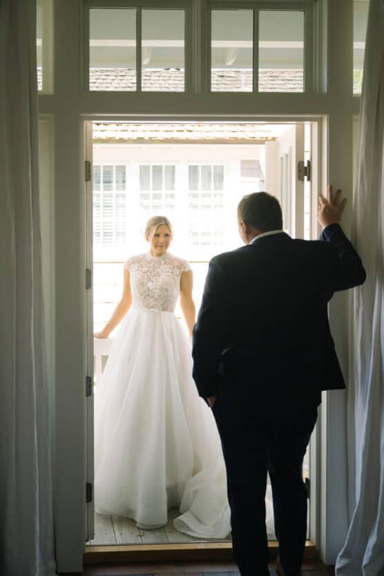 A bride in a white lace wedding dress stands on a porch, smiling, while a man in a suit stands in a doorway looking at her. Sunlight streams through large windows behind them. by Justin Salem Meyer