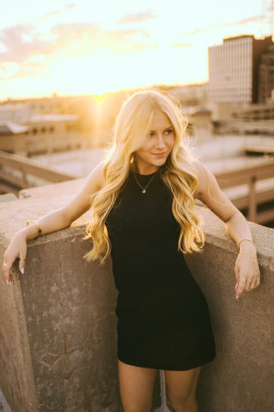 A young woman with long blonde hair in a black sleeveless dress leans against a concrete ledge on a rooftop at sunset, with golden sunlight and city buildings in the background. by Justin Salem Meyer