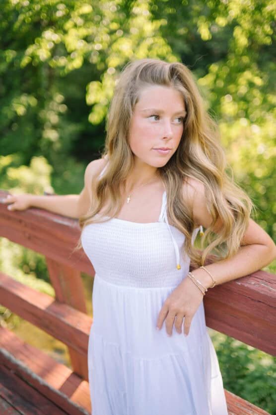 A young woman with long blonde hair in a white dress stands on a wooden bridge, surrounded by lush green foliage and sunlight. She looks thoughtfully into the distance, with one hand resting on the railing. by Justin Salem Meyer