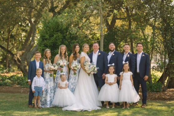 A wedding party poses outdoors. The bride and groom stand in the center, surrounded by bridesmaids in blue floral dresses, groomsmen in navy suits, and young flower girls and ring bearers in white outfits. Trees and greenery are in the background. by Justin Salem Meyer