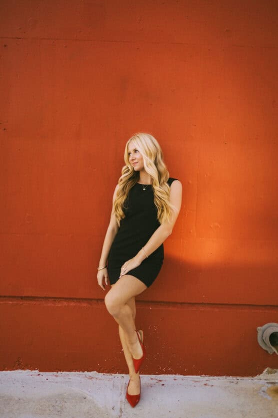 A woman with long blonde hair, wearing a black sleeveless dress and red heels, stands against a vivid red wall, looking to the side with one knee slightly bent. by Justin Salem Meyer