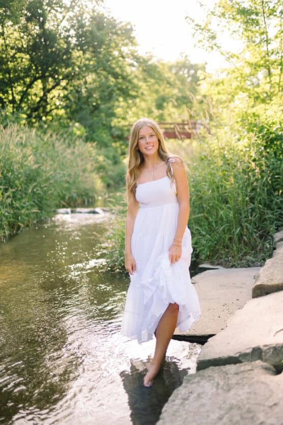 A young woman in a white dress stands barefoot on rocks in a shallow creek surrounded by lush green trees and grass, with sunlight streaming through the foliage. by Justin Salem Meyer