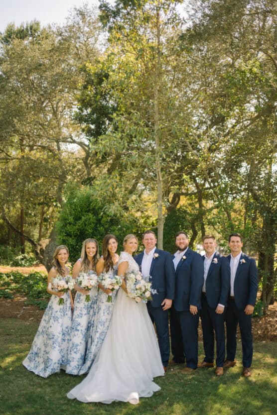 A wedding party poses outdoors in daylight. The bride in a white gown and four bridesmaids in blue floral dresses stand beside three groomsmen in navy suits, all smiling in front of green trees and sunlit foliage. by Justin Salem Meyer