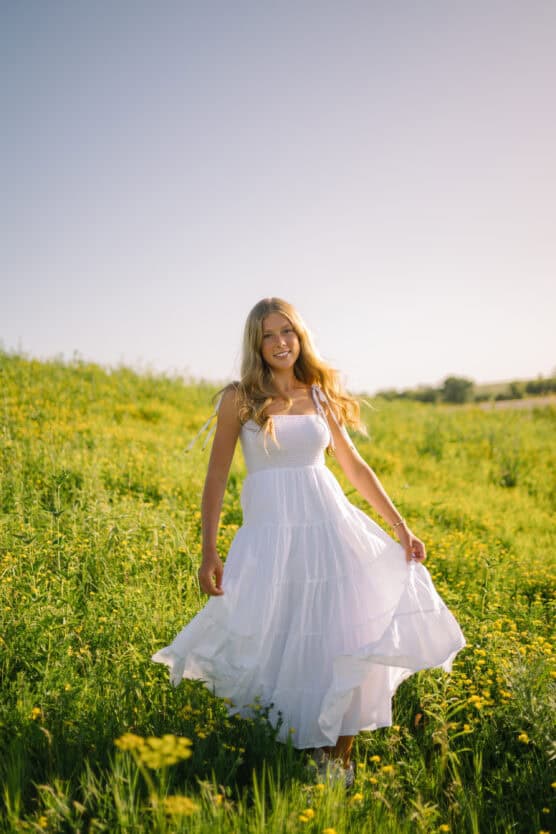 A woman in a flowing white dress stands in a sunlit field of green grass and yellow wildflowers, smiling and holding her dress as it sways. The sky is clear and bright, creating a peaceful, summery atmosphere. by Justin Salem Meyer