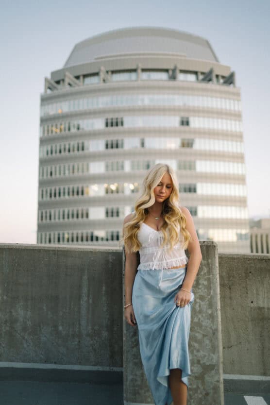 A woman with long blonde hair in a white top and light blue skirt stands in front of a tall, modern office building, leaning against a concrete barrier at sunset. by Justin Salem Meyer