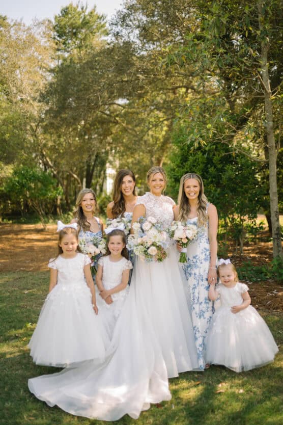 A bride in a white gown stands outdoors with four young flower girls in white dresses and two bridesmaids in floral dresses, all holding bouquets, surrounded by greenery and trees. by Justin Salem Meyer