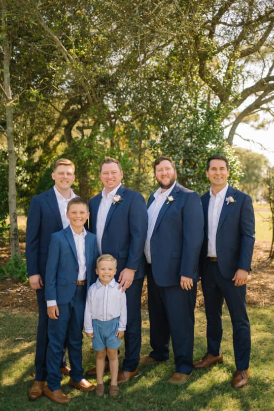 Five men and boys dressed in blue suits and white shirts stand together outdoors, smiling, with trees and sunlight in the background. The group includes three adults and two young boys. by Justin Salem Meyer