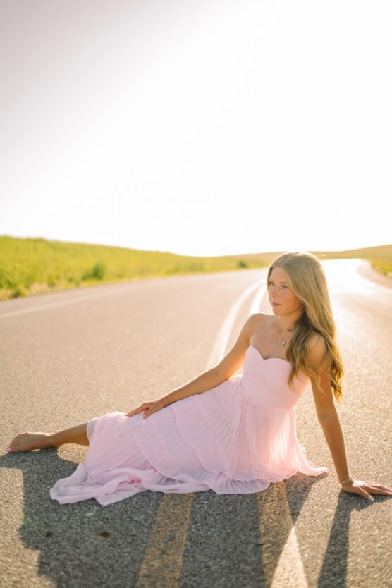 A young woman in a flowing light pink dress sits barefoot on a sunlit rural road, with green fields and bright sky in the background. by Justin Salem Meyer