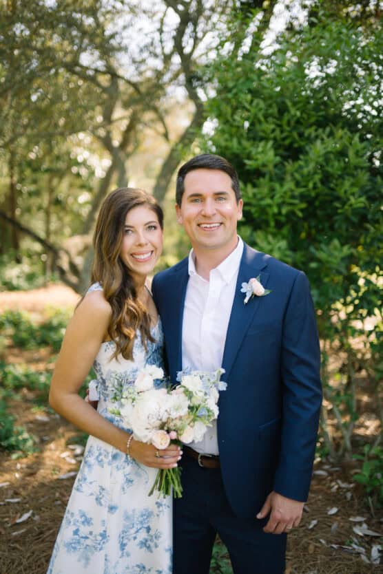 A smiling couple stands outdoors in a garden setting. The woman holds a bouquet of flowers and wears a white dress with blue floral patterns. The man wears a navy suit with a white shirt and floral boutonnière. by Justin Salem Meyer