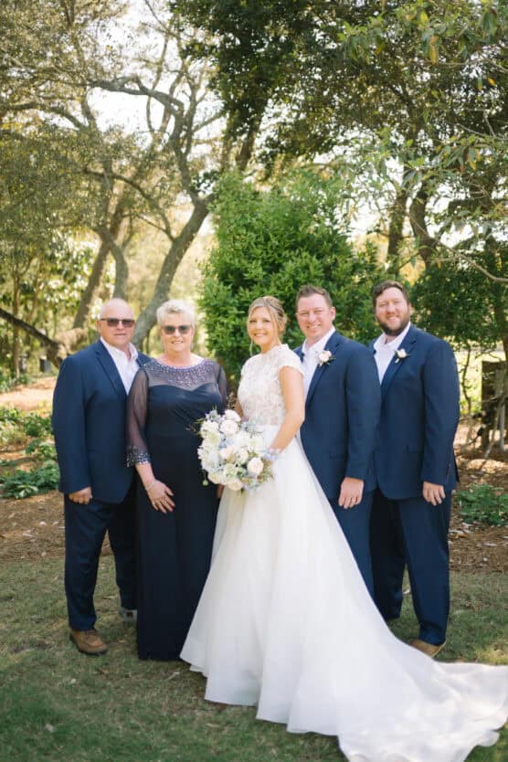 A bride in a white gown holds a bouquet, standing with four people—two men in navy suits, a woman in a navy dress, and another man in a navy suit—all smiling outdoors in a sunlit garden. by Justin Salem Meyer