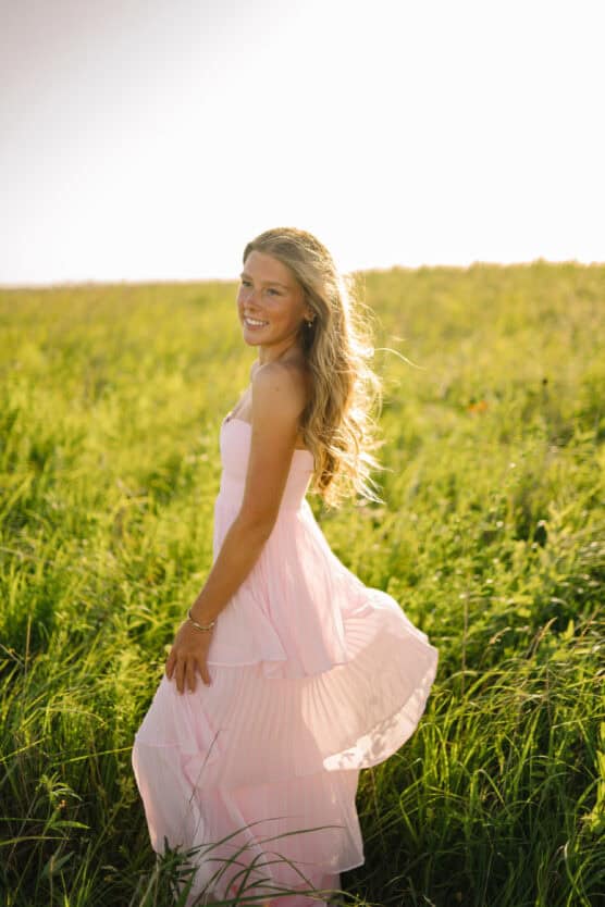 A young woman in a light pink dress stands in a sunlit field of tall green grass, smiling at the camera with her blonde hair flowing in the breeze. by Justin Salem Meyer