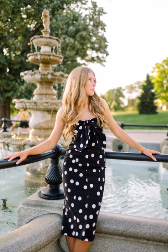 A woman with long blonde hair in a black and white polka dot dress stands by a fountain, holding onto a railing, with greenery and trees in the background on a sunny day. by Justin Salem Meyer