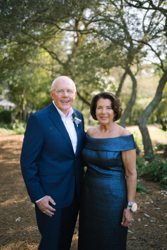 An older man in a blue suit and an older woman in an off-the-shoulder blue dress stand smiling together outdoors under trees, with sunlight filtering through the leaves. by Justin Salem Meyer