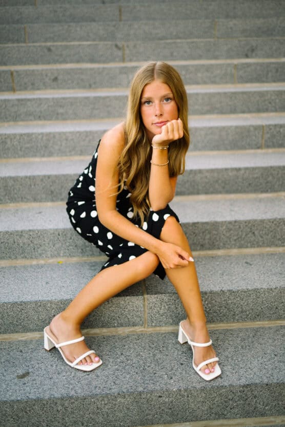 A young woman with long, wavy blonde hair sits on outdoor stone steps, wearing a black dress with white polka dots and white heeled sandals, resting her chin on her hand and looking at the camera. by Justin Salem Meyer