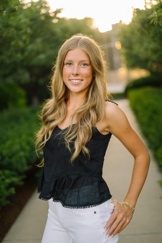 A young woman with long, wavy blonde hair stands on a sunlit path, wearing a black sleeveless top and white pants, smiling with one hand on her hip. Greenery lines the path in the background. by Justin Salem Meyer