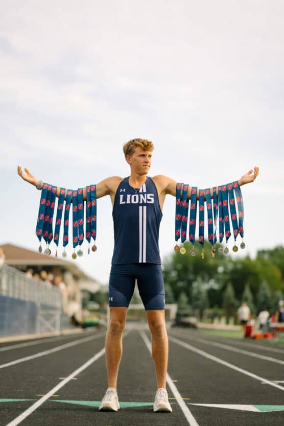 A young track athlete stands on a running track with arms outstretched, wearing a LIONS uniform and displaying multiple medals draped over both arms. The background shows trees and bleachers. by Justin Salem Meyer