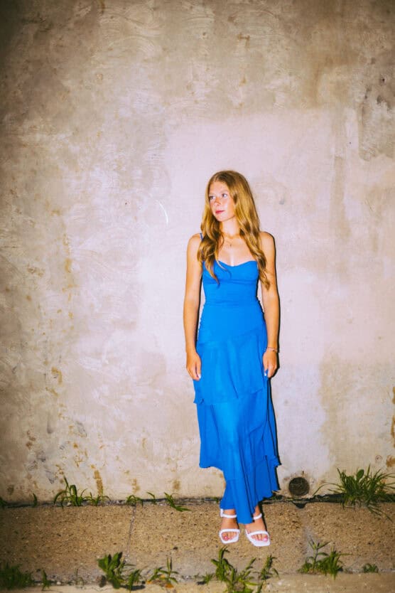 A woman in a bright blue dress and white sandals stands against a textured, beige wall on a concrete sidewalk with grass growing at the edges. She has long hair and is looking slightly to the side. by Justin Salem Meyer