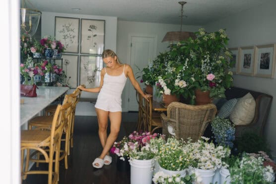 A woman in a white outfit stands in a room filled with various blooming flowers in pots and buckets, surrounded by wooden chairs and framed artwork on the walls. Natural light brightens the cozy, floral-filled space. by Justin Salem Meyer