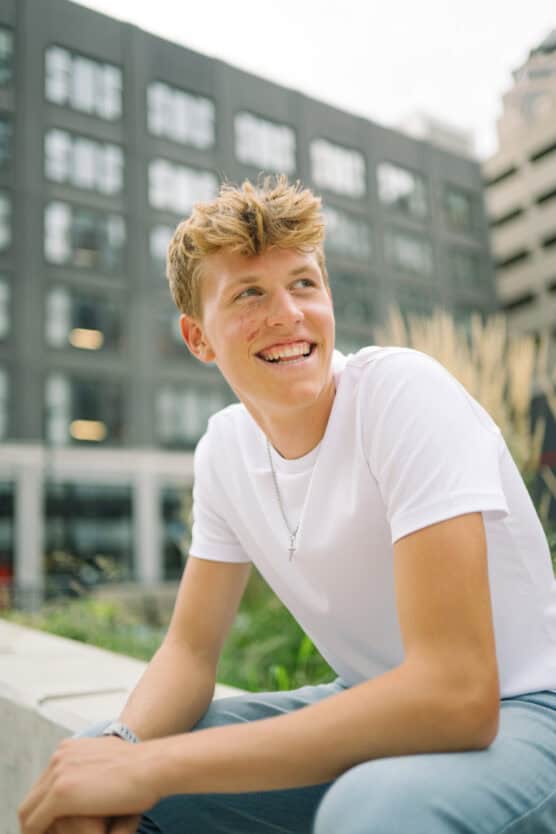 A young man with short blond hair, wearing a white t-shirt and silver necklace, sits outdoors on a bench, smiling and looking to the side, with modern buildings blurred in the background. by Justin Salem Meyer