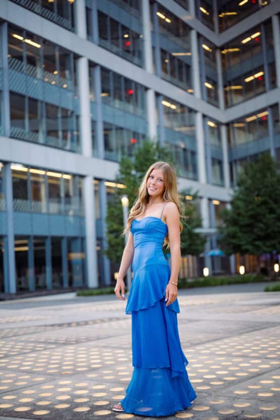 A young woman in a strapless blue dress stands outdoors, smiling in front of a modern glass building with trees and lights in the background. by Justin Salem Meyer