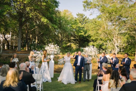 A bride and groom walk down the aisle outdoors, smiling and holding hands, while guests seated on white chairs applaud. Bridesmaids in floral dresses and groomsmen in blue suits stand nearby. Lush greenery surrounds the scene. by Justin Salem Meyer