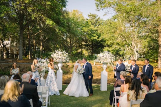 A bride and groom share a kiss at their outdoor wedding ceremony, surrounded by the bridal party, guests seated on white chairs, and elegant floral arrangements under trees in a sunlit garden setting. by Justin Salem Meyer