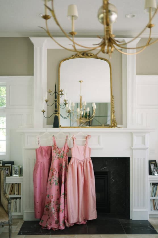 Three pink dresses hang from a white fireplace mantel beneath a large ornate mirror, with a chandelier reflected in the glass. The room is elegant, with framed photos on the mantel and classic white paneling. by Justin Salem Meyer
