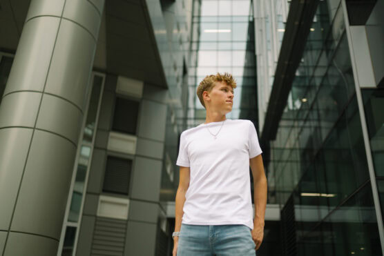 A young man in a white t-shirt and jeans stands outside a modern glass and metal building, looking to the side with a confident expression. The shot is taken from a low angle. by Justin Salem Meyer