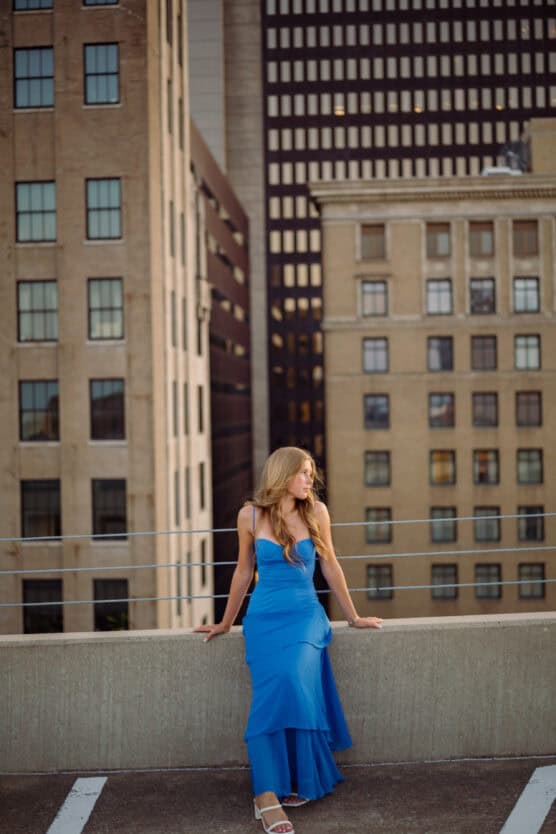 A woman in a long blue dress sits on a concrete ledge of a rooftop parking lot, with tall city buildings in the background at sunset. by Justin Salem Meyer