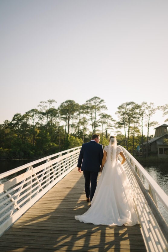A bride and groom walk hand-in-hand along a sunlit bridge, surrounded by trees and water, with the bride in a white dress and veil and the groom in a dark suit. by Justin Salem Meyer