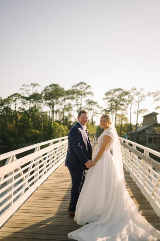 A bride and groom stand hand in hand on a sunlit wooden bridge, smiling back at the camera. The bride wears a white gown and veil, and the groom is dressed in a navy suit. Trees and a house are visible in the background. by Justin Salem Meyer